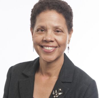 The photo features a smiling woman with short cropped hair on a white background. She is wearing a dark gray blazer with a fuchsia shirt underneath. She is wearing earrings and has a silver flower brooch on her left lapel. 
