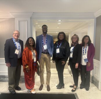 The photo features five people standing together in a row, smiling and all wearing white conference badges with blue lanyards around their necks. The location is a hallway with tan walls, white doors and paneling, bright lights, and white marble floors. An elegant bronze standing lamp is right behind and to the right of them, in the left hand edge of the frame. 