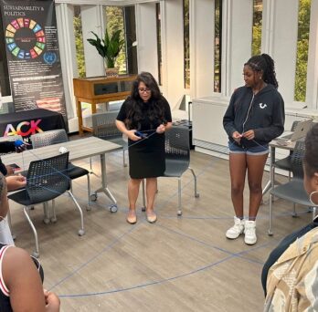 The photo features an instructor and several students standing in a circle in the middle of a UIC classroom. Each person is holding a part of a blue string and the string has many intersections as it runs between person to person. The instructor is holding a pair of scissors and preparing to cut the string. Posters about sustainability and politics as well as fresh water are propped up against the classroom walls. 