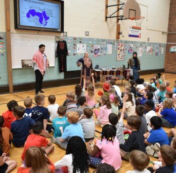 The photo features dozens of elementary school students sitting on a school gymnasium floor watching three university student presenters in traditional clothing from the Middle East and North Africa (MENA) regions. There are colorful tables set up behind the presenters with MENA regional artifacts and additional regional clothing hung up on the gymnasium wall. Above the presenters on the gymnasium wall is a projector screen with a PowerPoint slide showing the MENA region highlighted in purple. 