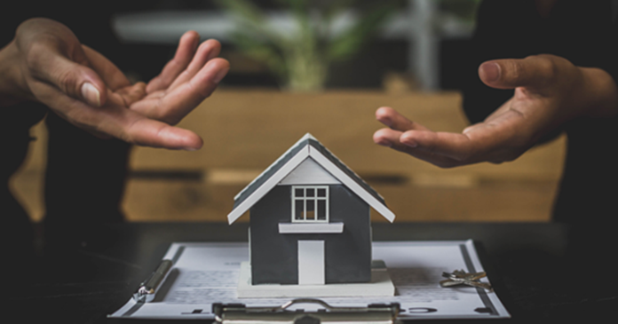 A small model of a house with a gray and white exterior sits on top of a clipboard with documents, possibly related to real estate or a property agreement. A set of keys is placed beside the clipboard. In the background, two hands are gesturing toward the house model, possibly indicating a discussion or negotiation. The scene is set on a dark table, with a blurred indoor environment in the background.