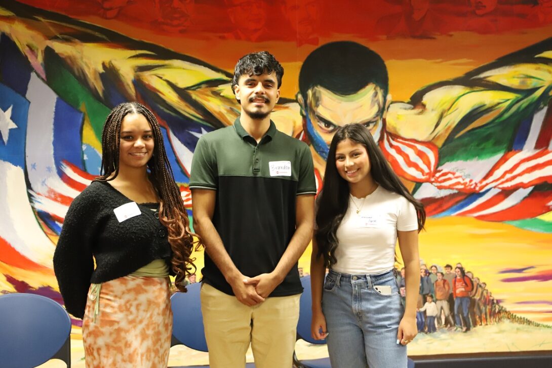 Three internship alumni stand next to each other on stage at the Diversity Community Engagement Program new student orientation in Fall 2023 at the Latino Cultural Center. The young woman on the left has long braided hair and wears an orange long skirt, the young man in the middle has a goatee and wears a black and green shirt and the young woman on the right wears light blue jeans and a white t-shirt. All three stand in front of the Awakening of the Americas mural and proudly smile right at the camera.