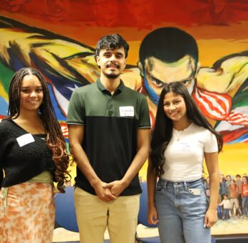 Three internship alumni stand next to each other on stage at the Diversity Community Engagement Program new student orientation in Fall 2023 at the Latino Cultural Center. The young woman on the left has long braided hair and wears an orange long skirt, the young man in the middle has a goatee and wears a black and green shirt and the young woman on the right wears light blue jeans and a white t-shirt. All three stand in front of the Awakening of the Americas mural and proudly smile right at the camera. 