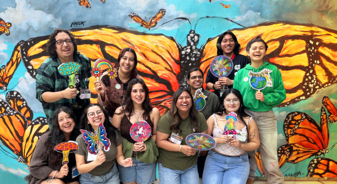 Ten smiling students pose in front of a large monarch butterfly mural, holding colorful hand-drawn paddle signs of mushrooms, butterflies, flowers and globes.