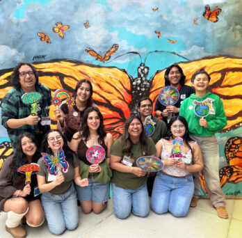 Ten smiling students pose in front of a large monarch butterfly mural, holding colorful hand-drawn paddle signs of mushrooms, butterflies, flowers and globes.